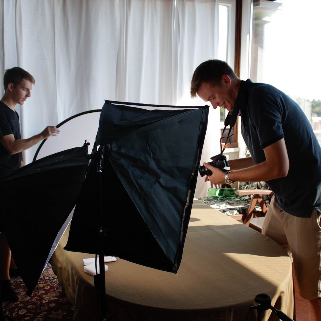 Two people setting up studio lighting and camera equipment in a room with large windows and curtains.