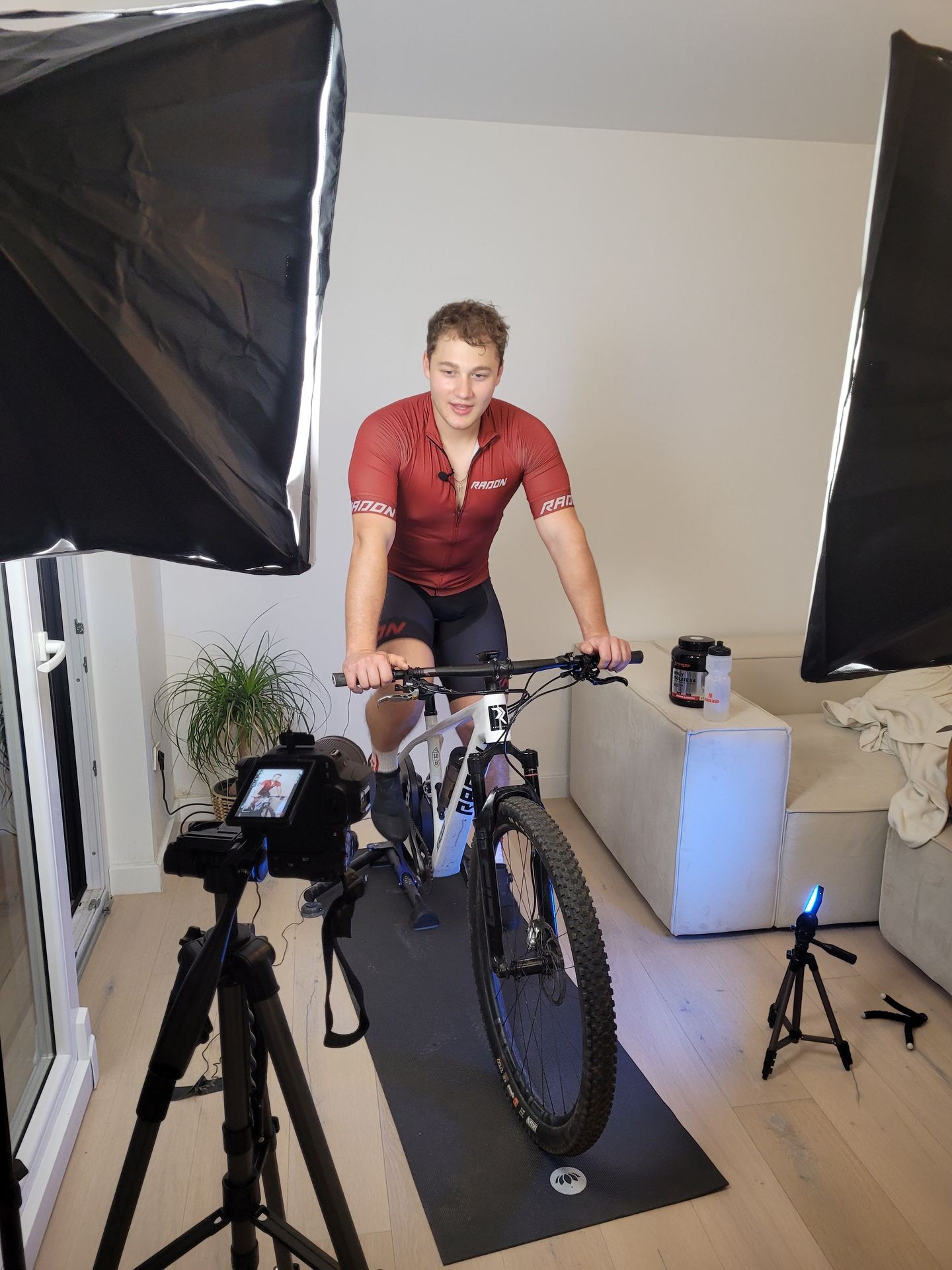 Man in a cycling outfit on a stationary bike indoors with professional lighting and camera setup around him.